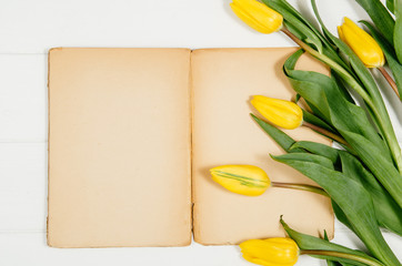 Background with yellow tulips, blank notepad on white wooden table top view. Creative woman's workspace concept with copy space. Product photograph taken from above with frame composition