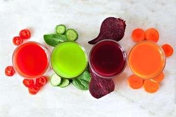 Four types of juice with scattered vegetables, above view over a white marble background