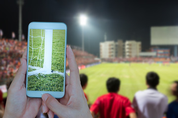 Hand holding smartphone with soccer screen in football stadium background.