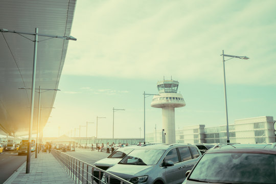 Parking lot and road near modern contemporary airport terminal in front of air traffic control tower, with many passengers and staff passing in distance, Barcelona, Spain