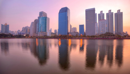 Fototapeta premium Beautiful city skyline of Bangkok at rosy dawn with lakeside skyscrapers and reflections ~ Morning view of modern buildings reflecting on smooth lake water in Benjakiti Park, Bangkok Thailand