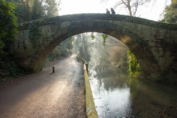 A couple walk across a solid stone hump back Victorian bridge as the golden wintry morning sun rays break through the trees and highlight the mist rising from the river