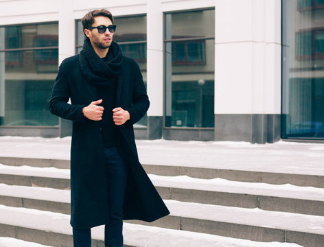 Elegant young handsome business man wearing suit and black coat posing against the background a business building. Fashion and Style. Streetstyle.