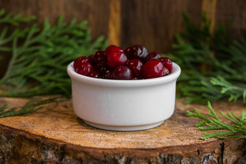 Pickled cranberries in a sweet syrup in a white bowl on the wooden background with the leaves of juniper