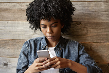 Beautiful young dark-skinned woman wearing trendy jeans jacket typing sms on her generic smart...