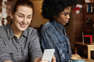 Gloomy African-American female having bored look, annoyed with her happy and cheerful Caucasian girlfriend, who is ignoring her, obsessed with mobile phone, messaging friends online at restaurant