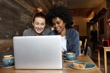 Modern technology, online communication, people and leisure concept. Two beautiful young females of different races sitting in front of open laptop, using free wireless internet connection at cafe