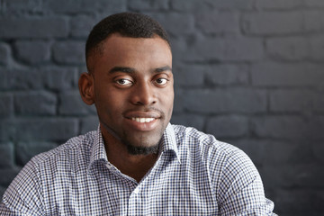 Close-up portrait of handsome young dark-skinned student or employee with small beard wearing formal checkered shirt looking and smiling at camera, sitting isolated against brick wall background