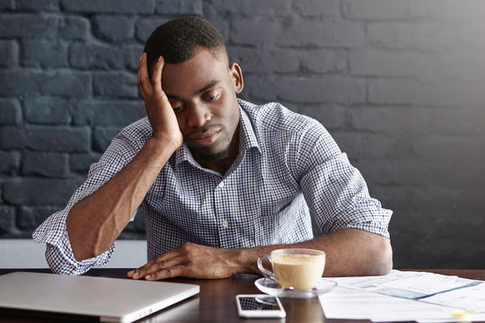 Frustrated tired young African-American employee touching his head, feeling absolutely exhausted because of overwork, calculating accounts, drinking another cup of coffee. Deadline and overwork