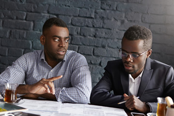 Two African-American businessmen discussing business plans at office: male in suit and glasses holding pen, studying papers on desk, his colleague having objections to him, raising his finger - obrazy, fototapety, plakaty