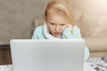 Stressed beautiful first-grade female pupil leaning on her elbow while doing homework using laptop pc, looking at screen with frustrated and unhappy expression, trying to complete tasks by herself