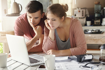 Serious man and woman sitting at kitchen table in front of open laptop computer, looking at screen...
