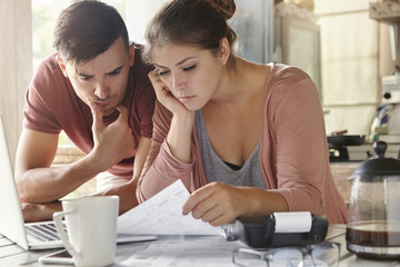 Young female and her unemployed husband with many debts doing paperwork together in kitchen,...