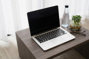 office table with blank screen on laptop, bottle of water, modern glasses, garden plant on glass vase on white drape background texture.