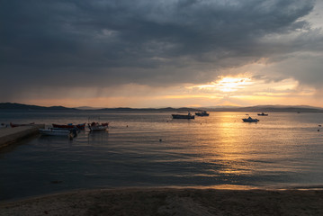 Fishing boats anchored in the bay at sunset with orange sun reflecting in the water and gathering storm clouds in the background