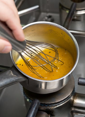 A metal mixing bowl full of fresh chicken stock being reduced over a flaming hob.The chicken stock is being whisked by hand and whisker.