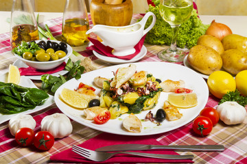 Grilled calamari on a white plate with boiled potatoes and chard, lemon, cherry tomatoes and olives, served next to fresh vegetables, sauce and glass of white wine.