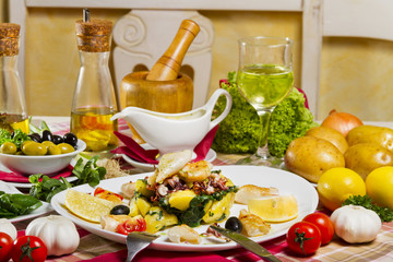 Grilled calamari on a white plate with boiled potatoes and chard, lemon, cherry tomatoes and olives, served next to fresh vegetables, sauce and glass of white wine.