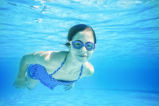 Smiling little girl swimming underwater in an outdoor swimming pool. Floating through the crystal blue water on a warm summer day