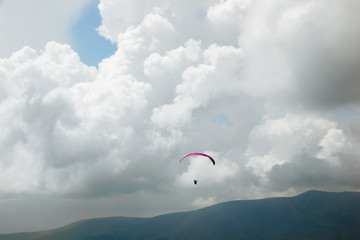 Paraglider flies among the clouds over green hills on a sunny day