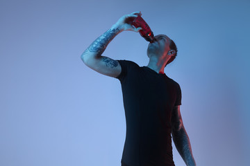 Fashion portrait of sport fit attractive man drinking water from bottle, tattooed hands, hipster look. Color flash studio light