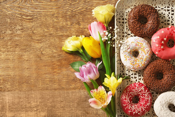 Tray with delicious donuts and colourful flowers on wooden background
