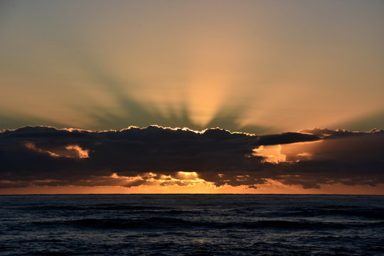 Beautiful sunrise cloudscape over ocean background. Sun rays beaming through picturesque clouds above sea. Blue sky with clouds, sea and sun on the horizon. Beautiful sunrise over the quiet calm sea.