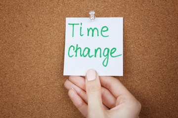 Female hand holding note with phrase TIME CHANGE on cork board background