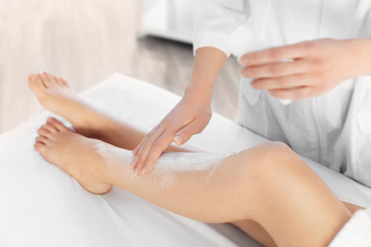 Beautician applying moisturizing cream on female legs in spa center