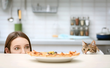 Young woman and cute cat looking at plate with tasty pizza in kitchen