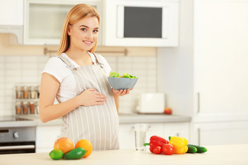 Young pregnant woman holding bowl with salad on kitchen