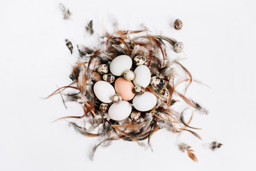 White, brown Easter eggs, quail eggs in nest decorated with feathers on white background. Flat lay, top view. Traditional spring concept.