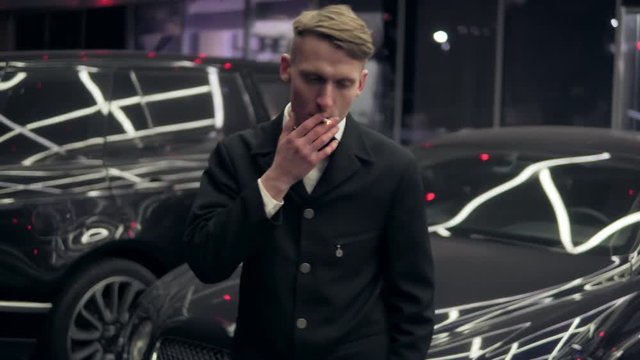 Portrait of a young man in a black suit with a bow-tie smoking a cigarette by the black car at night.