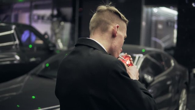 Young man in a black suit with a bow-tie smoking a cigarette by the black car at night.