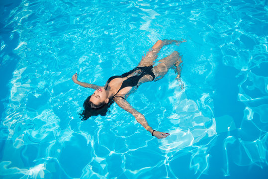 Attractive Woman In A Black Bathing Suit Floating On Her Back In The Swimming Pool And Relaxing, With Copy Space