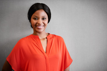 Portrait of black businesswoman smiling at camera while wearing a bright orange blouse while standing against a textured grey wall.