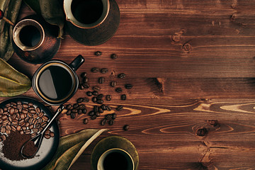 Hot coffee in black cup and several turkish pots cezve with beans, dry leaves with copy space on brown old wooden board background, top view. Rustic style.