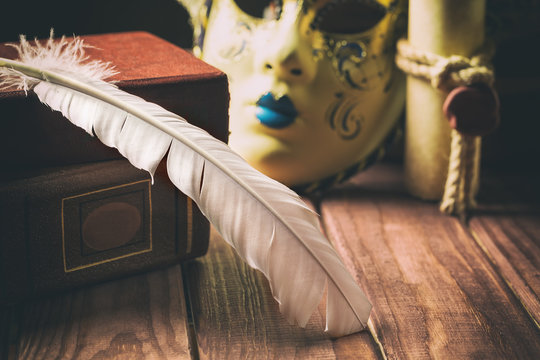Literature concept. Feather on book near venetian mask and old scroll on wooden background