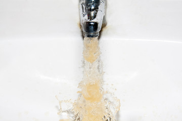 Dirty brown running water falling into a white sink from tap - obrazy, fototapety, plakaty