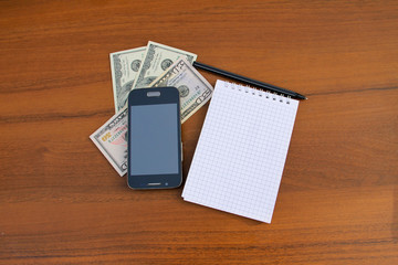 Notepad with pen, smartphone and dollar cash on wooden desk