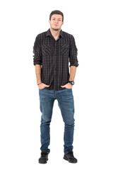 Smirking young casual man wearing jeans and plaid shirt with hands in pockets. Full body length portrait isolated over white background.