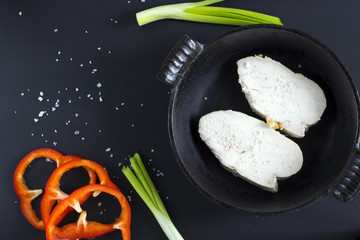 Baked white meat in iron pan with sea salt, red paprika, green onion, leafy green salad and cayenne pepper on black background