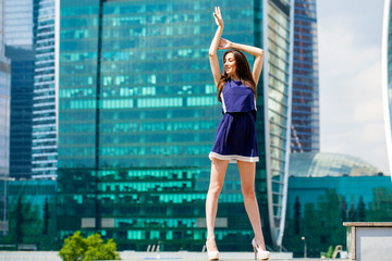 Fototapeta na wymiar Young woman in a blue dress is stretching near skyscrapers