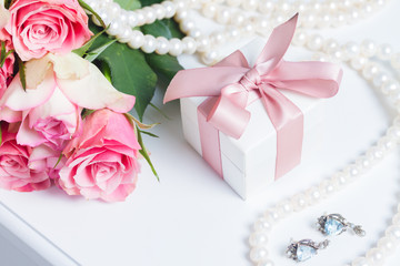 Present box with pink ribbon with jewellery and roses on white table