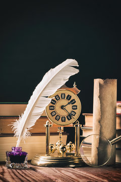 Literature concept. Inkstand with feather near magnifying glass, old scroll and vintage clock against dark background with books.