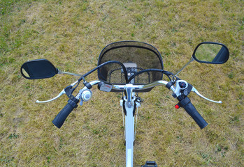 Fototapeta na wymiar Electric bicycle rudder or helm in the park on sunny summer day. Shot from above. The view of e bike steering wheel with bell, brakes, speed switch, and power battery control device and mirrors.