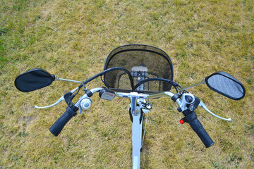 Fototapeta na wymiar Electric bicycle rudder or helm in the park on sunny summer day. Shot from above. The view of e bike steering wheel with bell, brakes, speed switch, and power battery control device and mirrors.