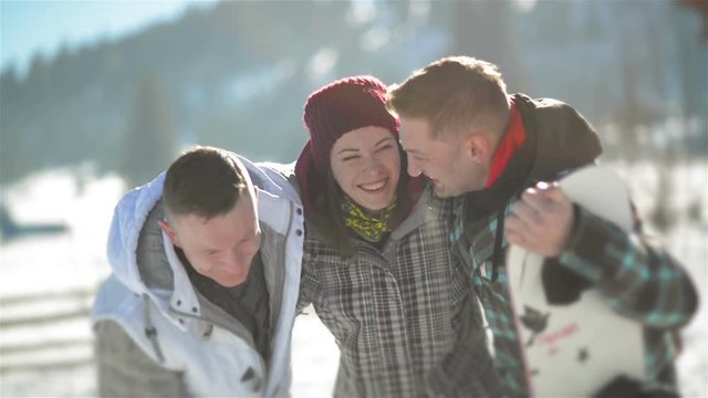 Two Men and Woman are Hugging Each Other Standing Outside During Warm Winter Day in the Mountains. One Man is Holding White Snowboard. Vacation Concept.