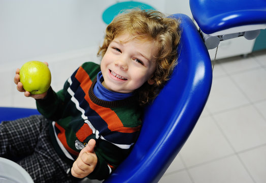 small child with curly hair in a striped sweater, sitting in the dental chair and holding a green apple. Examination of the teeth at the dentist.