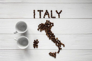 Map of the Italy made of roasted coffee beans laying on white wooden textured background with two coffee cups. Space for text